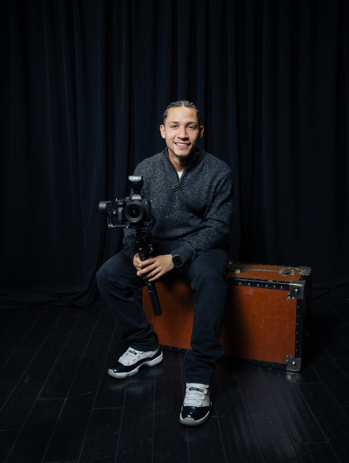 Ray Alameda seated on a vintage trunk in front of a dark curtain, smiling and holding his Sony camera on a gimbal.